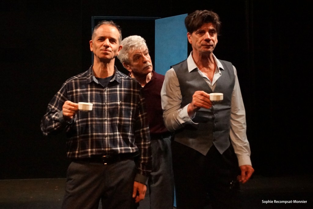 Trois hommes sur scène, chacun tenant une tasse, avec une porte bleue en arrière-plan.